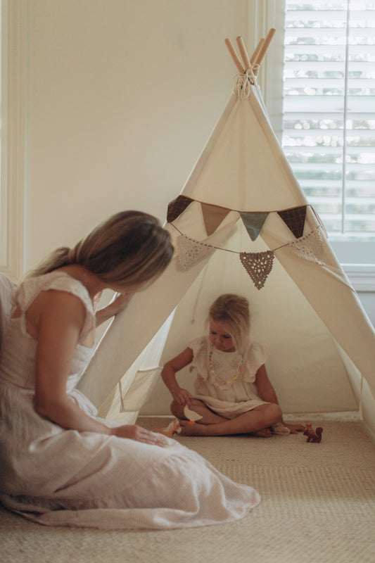 A mother sitting with her three-year-old child inside a fabric teepee, sharing quiet pretend play in a calm, cozy room.