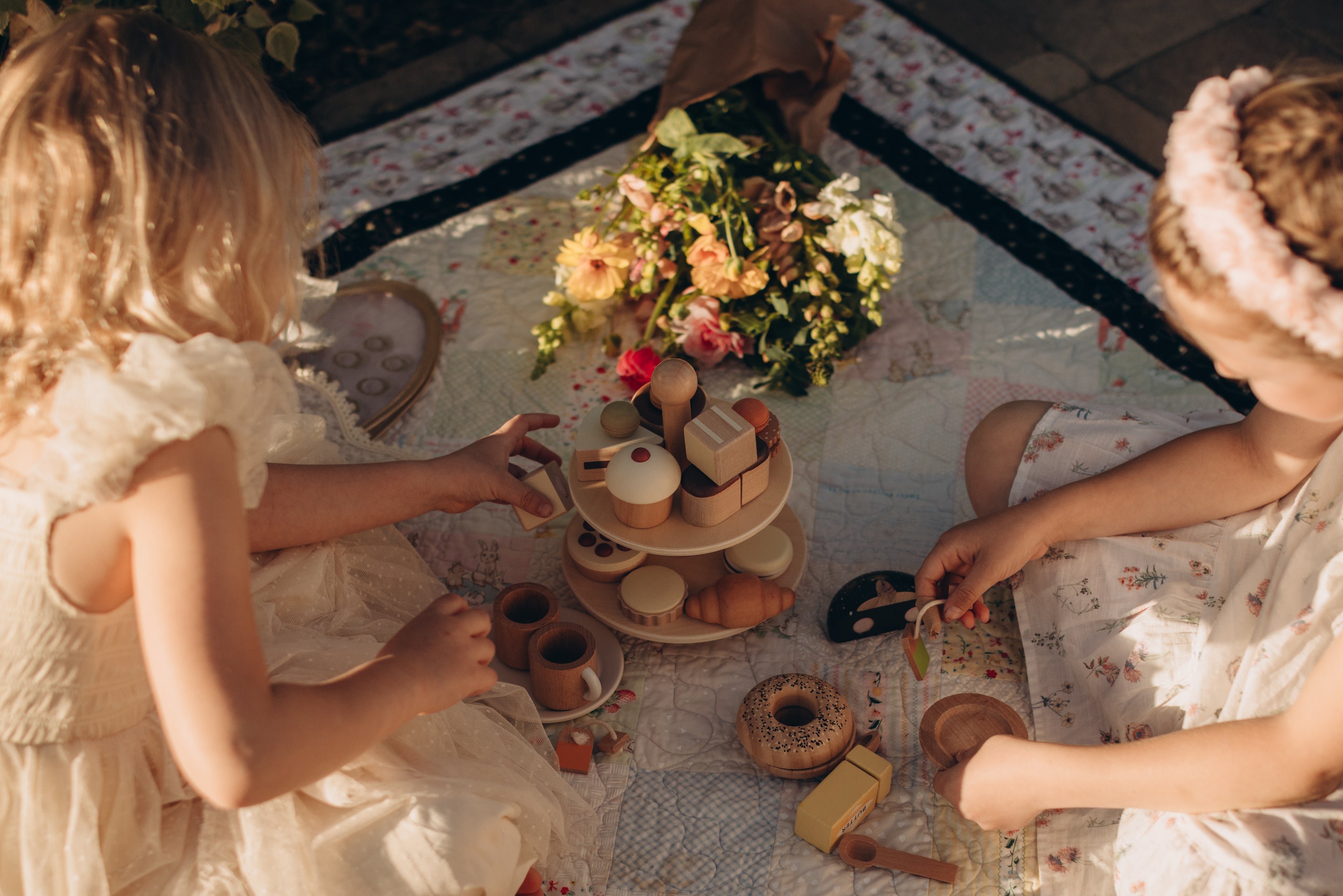 Two children in floral dresses sitting on a patterned rug with a small table of food and flowers.