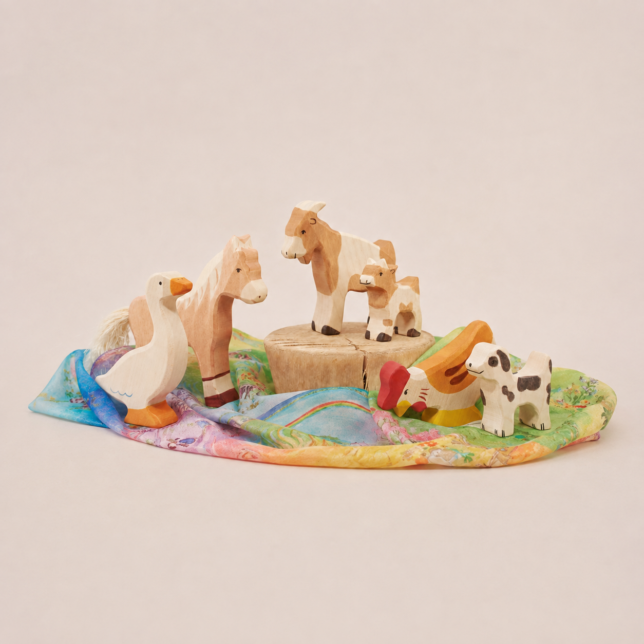 Wooden farm animal toys arranged on a colorful Playsilk, featuring a horse, goats, goose, chicken, and dog.