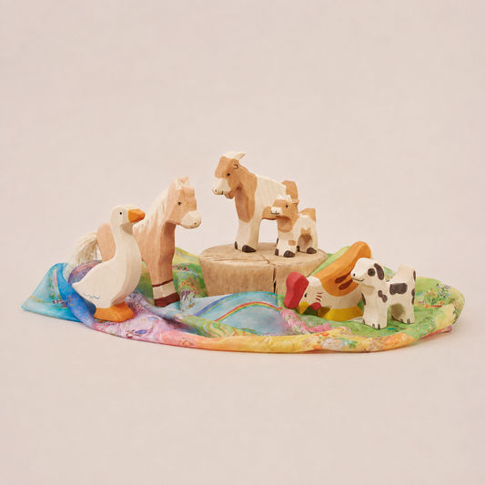 Wooden farm animal toys arranged on a colorful Playsilk, featuring a horse, goats, goose, chicken, and dog.