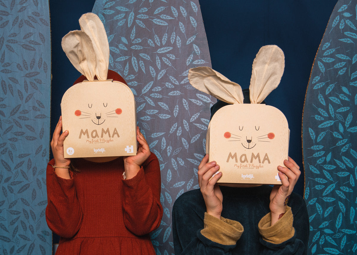 Two people holding up cardboard boxes with bunny ears and the word 'MAMA' on the front, posed in front of a blue patterned backdrop.