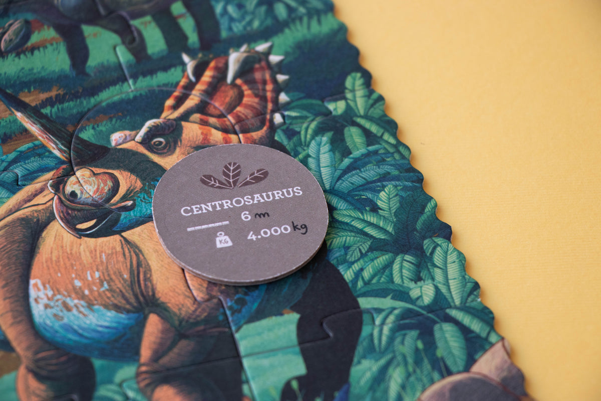 A close-up of a dinosaur-themed fabric or wallpaper sample with a scalloped edge, featuring an illustrated brown dinosaur among green foliage. A round gray tag reading 'CENTROSAURUS 6 m 4.
