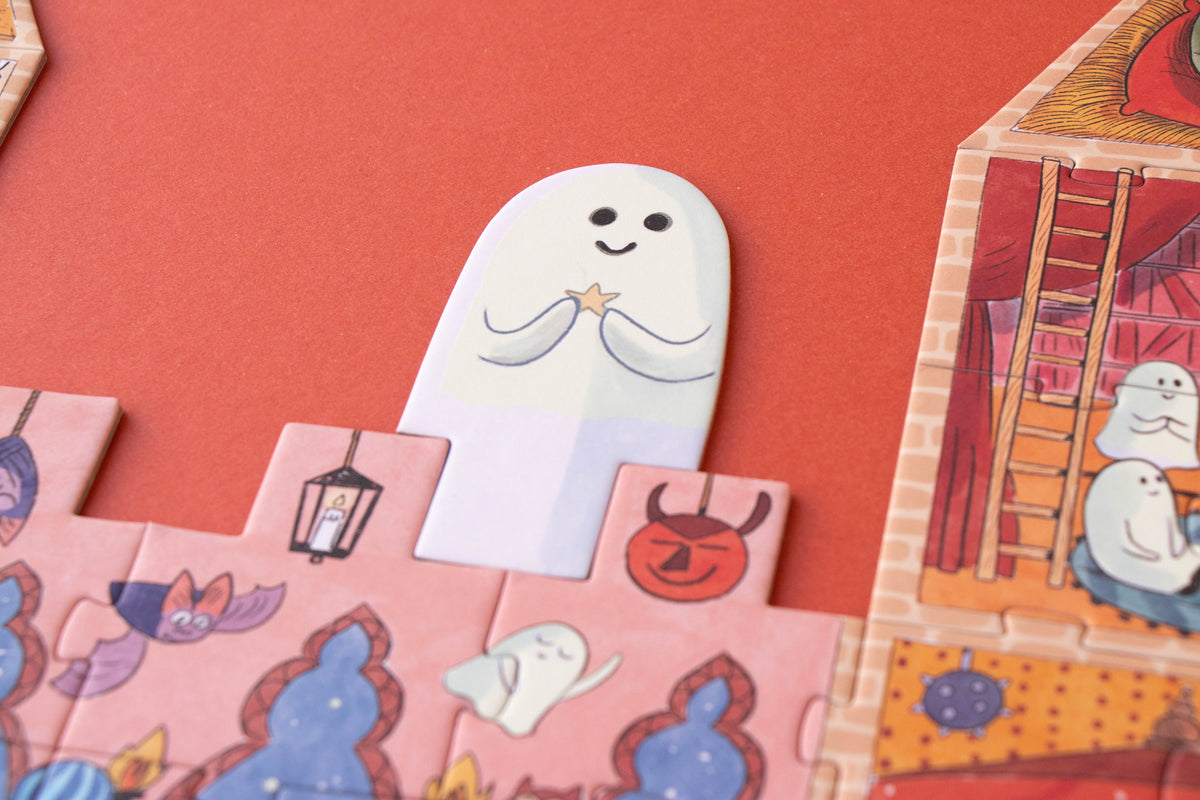 A close-up of a children's puzzle or card game featuring cartoon ghost illustrations. A white ghost-shaped piece is centered on a coral-red background, with pink and orange illustrated puzzle pieces around it showing Halloween-themed designs like a lantern, devil face, and house interior.