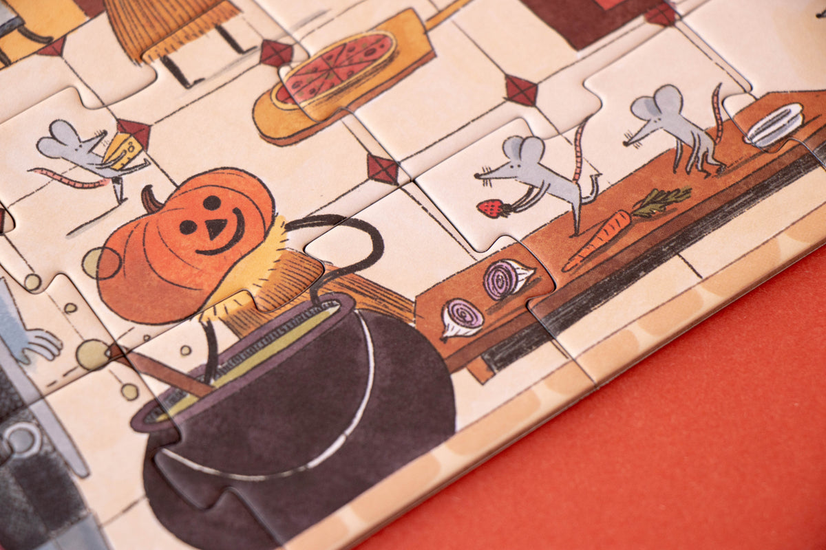 A close-up of a wooden jigsaw puzzle featuring a whimsical Halloween kitchen scene with a smiling orange pumpkin, a black cauldron, gray mice, and food items on a beige tile background, set against a solid orange surface.