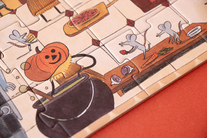 A close-up of a wooden jigsaw puzzle featuring a whimsical Halloween kitchen scene with a smiling orange pumpkin, a black cauldron, gray mice, and food items on a beige tile background, set against a solid orange surface.