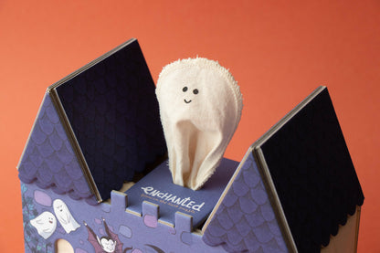 A plush white ghost finger puppet with a smiling face stands inside a purple cardboard house-shaped box labeled 'Enchanted' on an orange background.