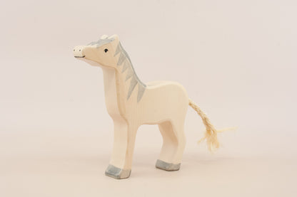 Hand-carved wooden toy horse in a natural light wood finish with painted gray details on the mane, hooves, and face, and a braided straw tail, shown against a plain light beige background.