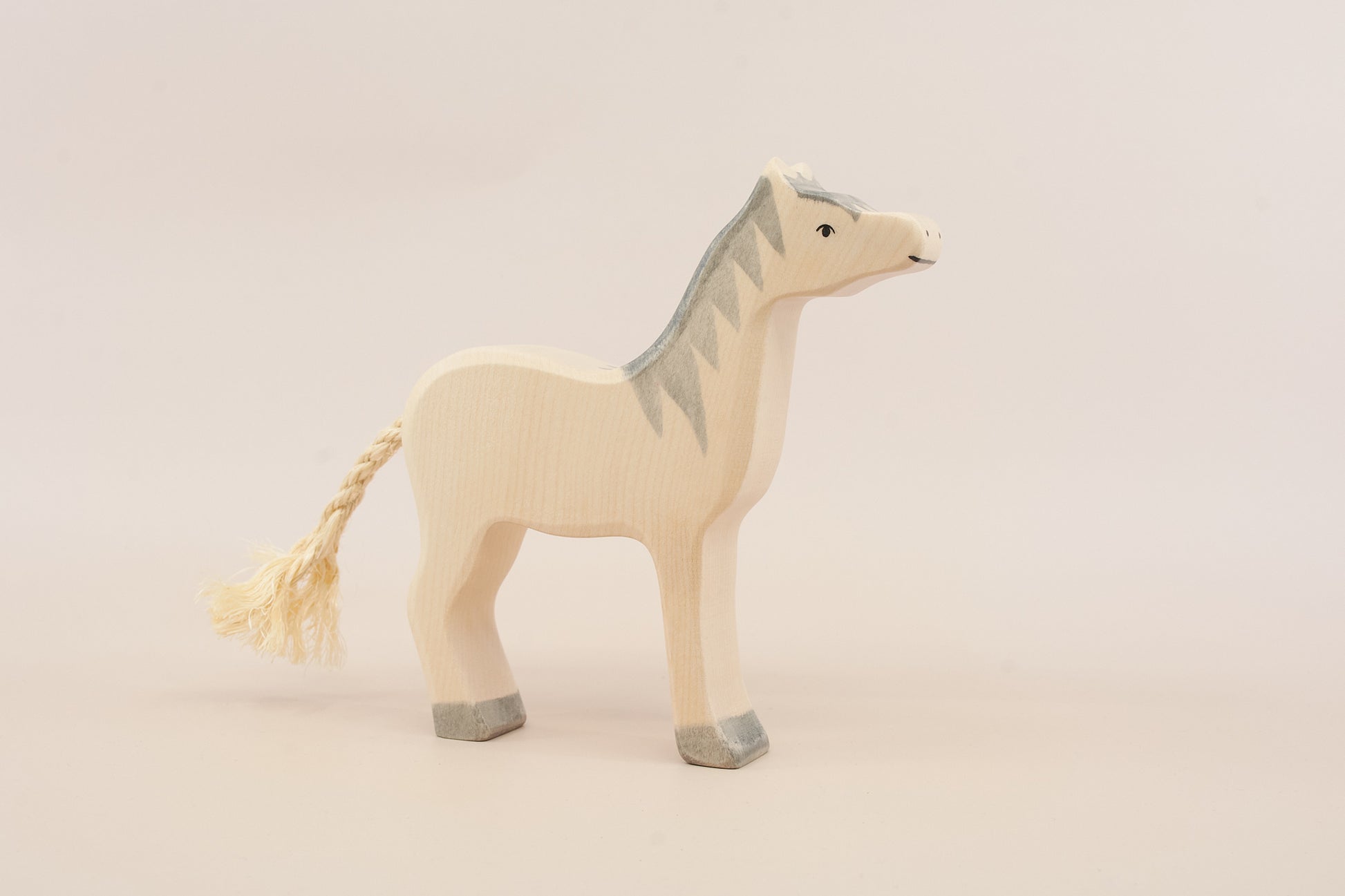 A minimalist wooden toy horse figurine with a cream-colored body, gray painted mane and hooves, and a braided cream tail, set against a solid light beige background.