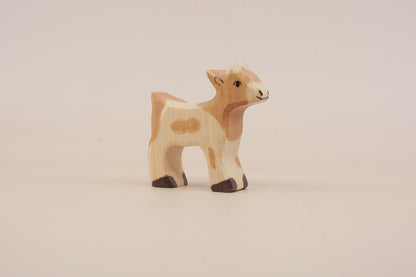 A small handcrafted wooden toy calf figurine with natural wood grain, painted with light brown and cream spots, standing on a plain light beige background.